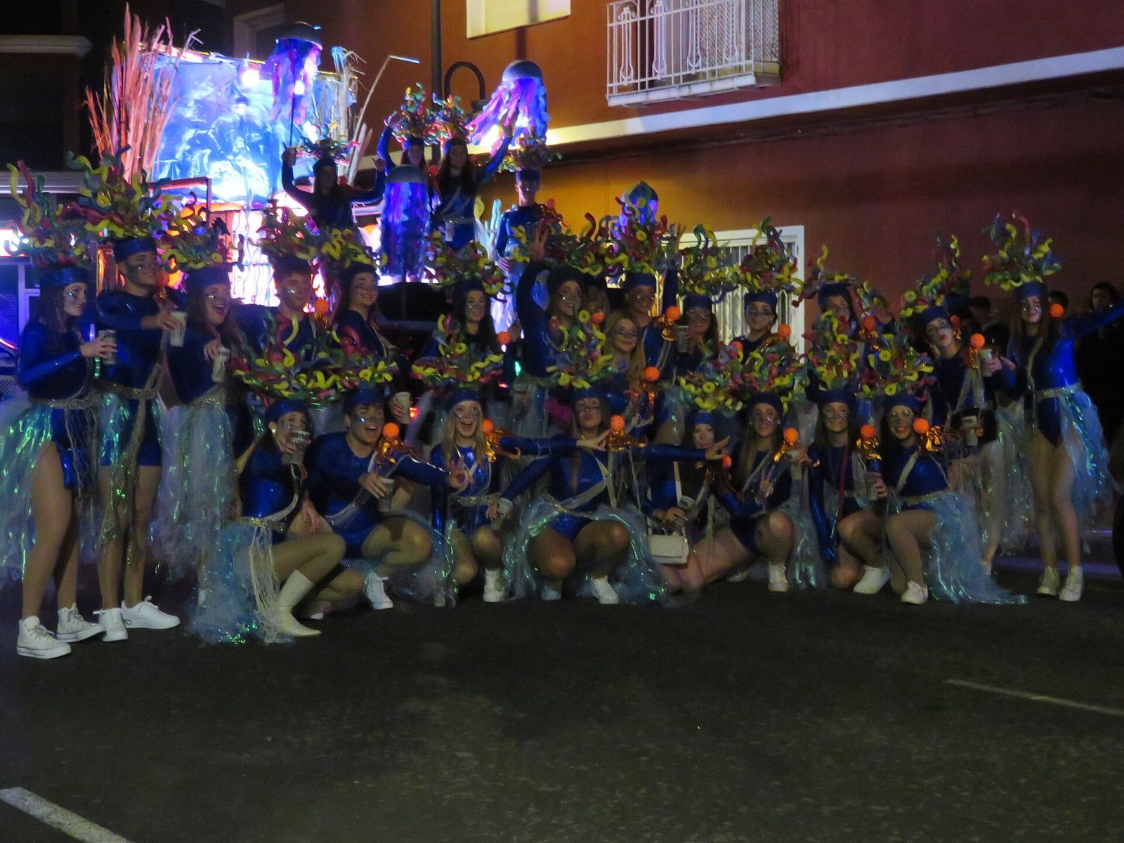 Comparsa con disfraces artesanales azules y tocados de colores posando ante su carroza en el desfile nocturno de Carnaval de Alcalà de Xivert